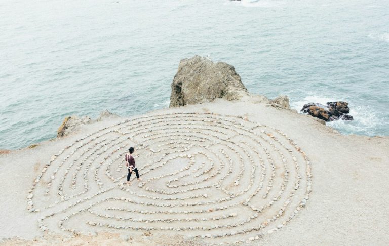 Erfahre mehr auf der Seite Begleitung Person geht durch einen Steinkreislauf am Meer. Felsen und Wasser im Hintergrund.