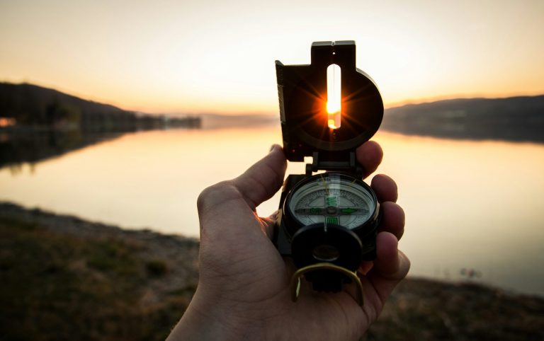 Erfahre mehr über Methoden auf der Seite Begleitung Eine Hand hält einen Kompass vor einem Sonnenuntergang am Wasser.